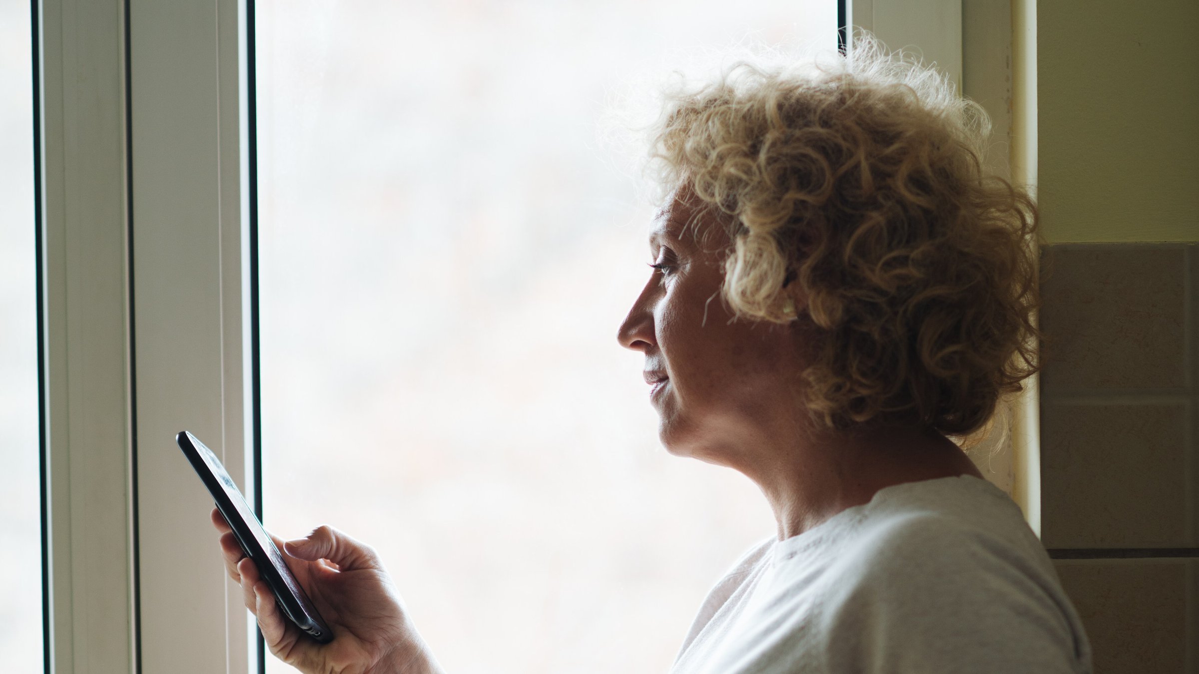 Een oudere vrouw bekijkt haar smartphone bij het raam
