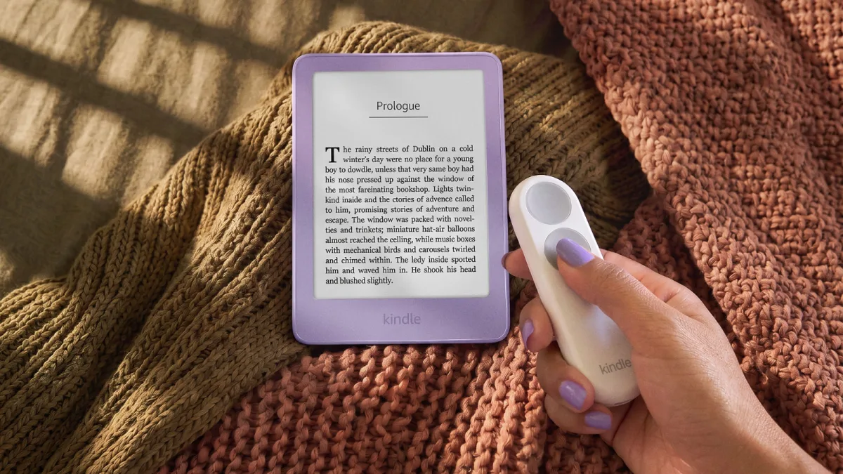 De nieuwe Kindle in de kleur Ube op een deken, met de Kindle Click-afstandsbediening in een hand